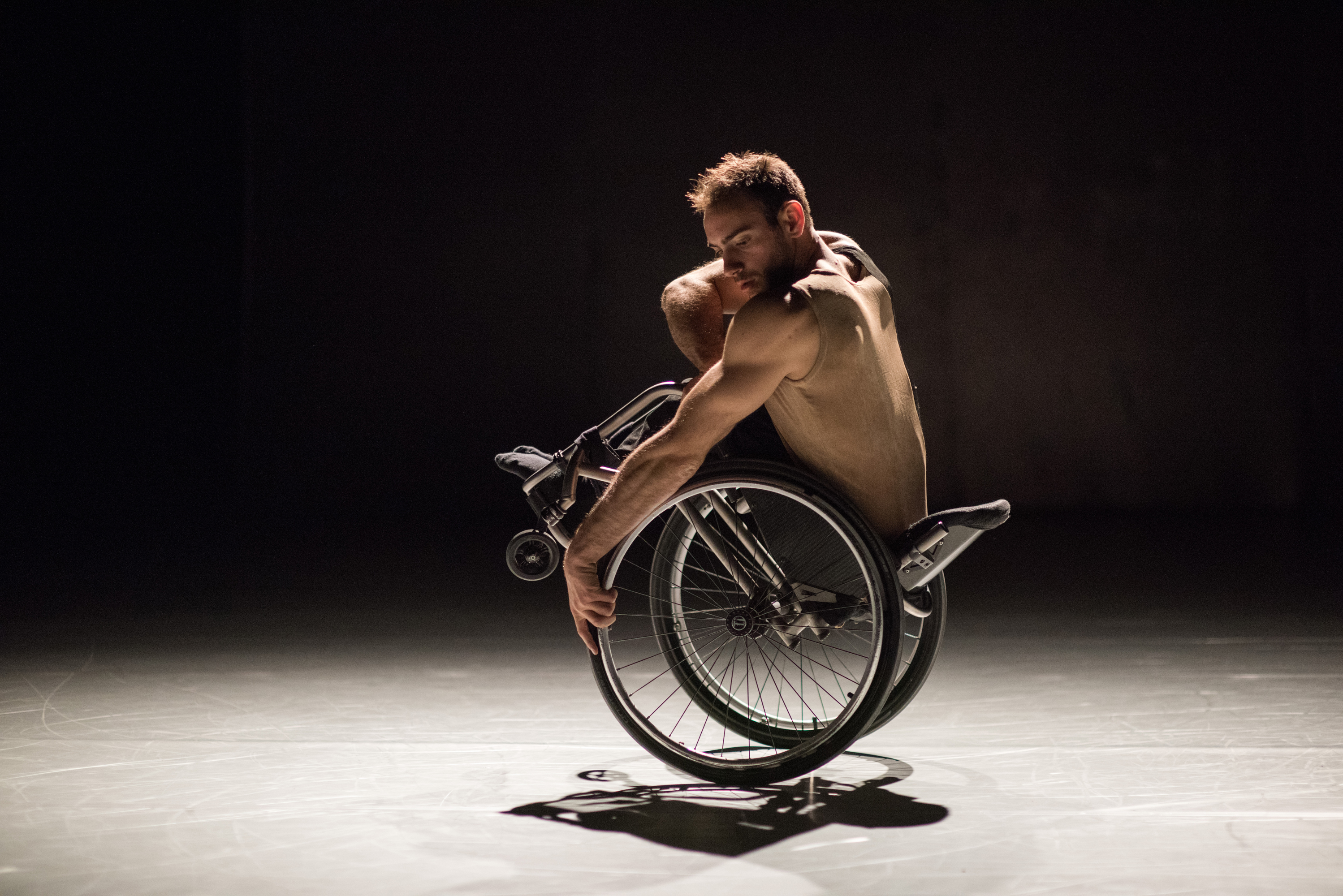 Dansaren Joel Brown på scen i verket "Beheld" av Alexander Whitley. Joel starka armar är framträdande och han tittar koncentrereat ner i scengolvet samtidigt som han balanserar bakåt med sin rullstol bakåt på sin rullstol.