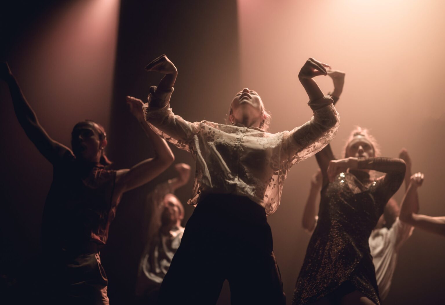 A group of 5 dancers pose together, with one female dancer in the foreground and the other 4 dancers positioned at different points behind her. The dancer in the foreground is leaning backwards slightly and has her arms out to each side, bent at the elbow, and her hands curve outwards in a pinching motion. Her head is tilted upwards, her facial expressions are relaxed and her mouth is ajar. Behind her, the other dancers take up similar positions, with one of the dancers holding one arm above her head instead of both. The dancers are wearing an assortment of different colourful shirts, trousers and dresses, with the female dancer in the foreground wearing a flowing, white shirt.